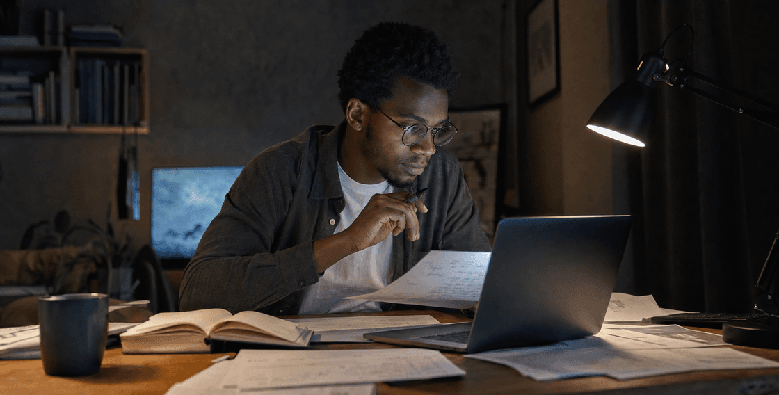 Person carefully reviewing laptop output against handwritten notes and printed pages in a dim workspace